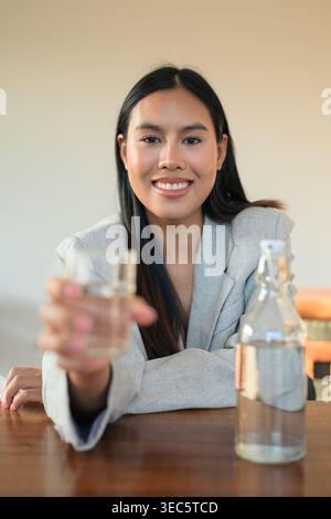 Freundliche thailändische Profi-Frau, die Wasser in einem Blazer am Schreibtisch anbietet. Dieses hochwertige, unveränderte Foto zeigt eine warme menschliche Mutter Stockfoto