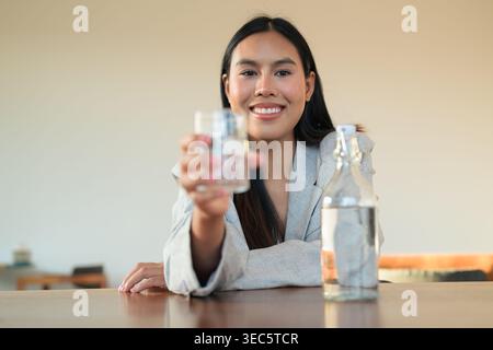 Fröhliche thailändische Profi-Frau, die Wasser in einem Blazer am Schreibtisch anbietet. Dieses hochwertige, unveränderte Foto zeigt eine warme menschliche Mutter Stockfoto
