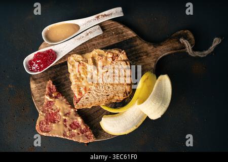 Eine Portion gegrillter Erdnussbutter- und Gelee-Sandwiches, von denen eine Hälfte bereits gegessen wurde, wird auf einem Holzbrett neben einer geschälten Banane und spo präsentiert Stockfoto