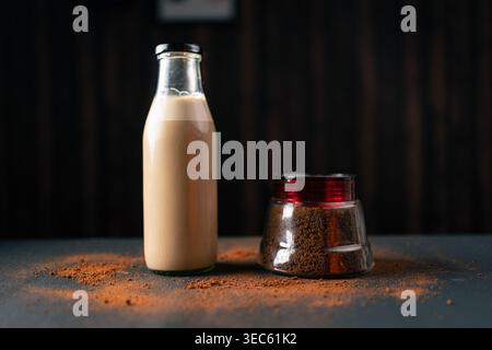 Eine klare Glasflasche mit cremigem Eiskaffee steht neben einem kleinen Glas Kaffeesatz mit verstreutem Pulver auf einer dunklen Oberfläche, was eine Zubereitung andeutet Stockfoto
