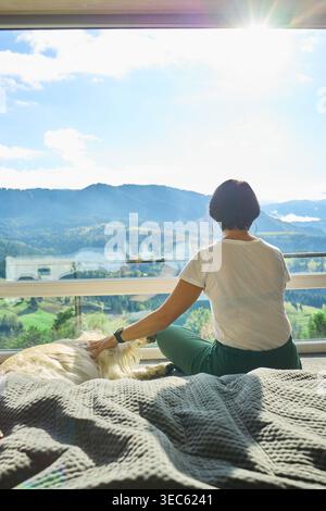 Frau sitzt auf einem Sofa mit Hund vor einem großen Fenster mit Blick auf die Berge bei Tageslicht Stockfoto