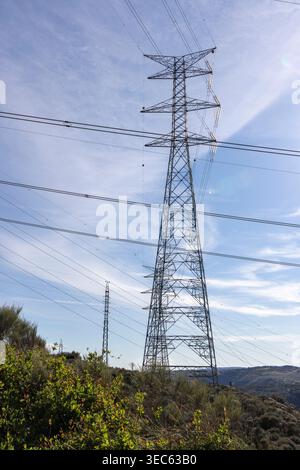 Gitterstahlleitungen mit Hochspannungsleitungen über eine grüne Landschaft unter blauem Himmel, symbolisiert die Energieinfrastruktur Stockfoto