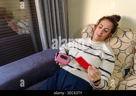 Frau, die sich auf einem Sofa entspannt, einen tragbaren Kassettenspieler und ein Kassettenband hält, Musik mit Kopfhörern hört und Nostalgie annimmt Stockfoto