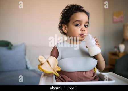 Porträt eines Kleinkindes, das Milch aus der Flasche trinkt, während er Banane im Hochstuhl hält, zeigt Selbstfütterung Stockfoto