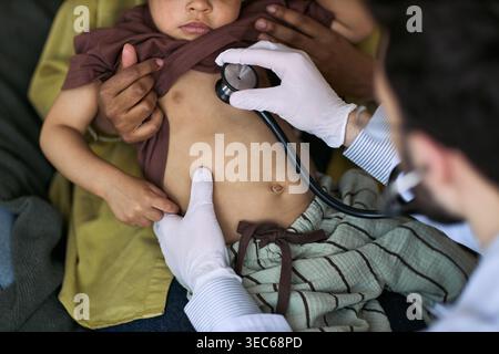 Nahaufnahme eines Arztes, der Kleinkinderbrust mit Stethoskop untersucht, während der Elternteil das Kind während der Untersuchung hält Stockfoto