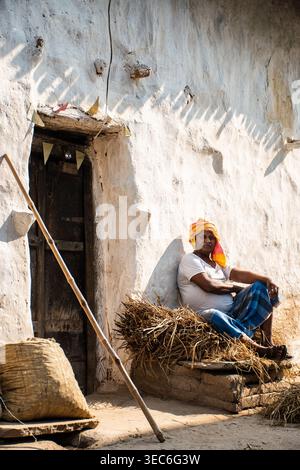 Ein Mann mit einem leuchtend gelben Turban sitzt nachdenklich vor einem rustikalen, weiß getünchten Haus in Bihar, Indien, getaucht in warmes Sonnenlicht, und blickt in die Th Stockfoto