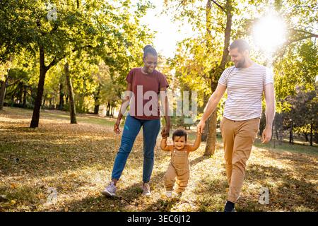 Eine fröhliche Familie genießt einen gemütlichen Spaziergang durch einen sonnigen Park voller Herbstfarben. Ein glückliches Kind wird spielerisch von seinen Eltern hochgezogen und strahlt Liebe und Wärme aus. Stockfoto
