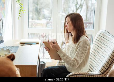 Frau, die Pappkarton öffnet, während sie am Schreibtisch sitzt, mit Hund im Hintergrund Stockfoto