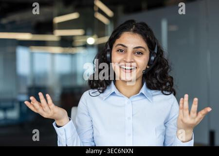 Lächelnde junge indische Geschäftsfrau aus dem Nahen Osten mit Headset, die während eines Videogesprächs im Büro spricht. Diverse multikulturelle weibliche Unterstützungsspezialistin an Stockfoto
