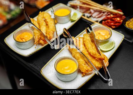 Goldene knusprige Garnelen auf rechteckigen Tellern, begleitet von kleinen Schüsseln mit cremiger Sauce und Limette. Stockfoto