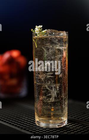 Ein hohes Glas kristallklares Getränk mit Eiswürfeln und zarten Blumendekorationen vor dunklem Studiohintergrund. Stockfoto