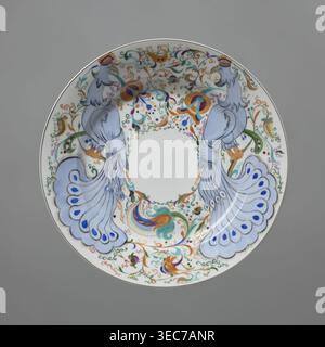 Platte, Porzellan; Fabrik benannt nach Michail Wassiljewitsch Lomonossow 1918–. Teller mit farbenfroher Dekoration, verteilt in einem breiten Band um den Rand – zwei stilisierte blaue Pfauen zwischen Pflanzenrollen und Orchideenblüten; Mitte links undekoriert. Schüssel; Porzellan; 1920; Leningrad, Russland. Höhe 4 cm; Durchmesser 24,8 cm., Eine runde dekorative Platte mit weißem Hintergrund mit einem symmetrischen Design von zwei stilisierten Vögeln, die sich zueinander gegenüberstehen, jeweils in blassblau mit langen fließenden Schwanzfedern mit dunkleren blauen tropfenförmigen Akzenten und goldenen Schnäbeln mit kleinen kronenartigen Wappen; die Stockfoto