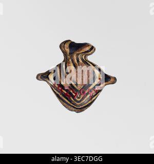 Amulett in Form eines Herzens, aus schwarzem und braunem Glas, verziert mit parallelen Linien, die auf der Vorder- und Rückseite einen Zickzack bilden, in Beige und weiß. Das Amulett ist vertikal durchbohrt, das Loch ist oben mit einem beigefarbenen Ring eingerahmt, und an den Seiten befinden sich Restgriffe. 1550–1295 v. Chr.; Neues Königreich, 18. Dynastie, Ägypten. Amulett; Höhe 1,6 cm, Breite 1,7 cm., Ein kleines Objekt mit organischer, unregelmäßiger Form sitzt vor einem einfachen hellgrauen Hintergrund; das Objekt hat mehrschichtige, fließende Streifen in Schwarz- und Braun-/Goldtönen mit Bereichen in Braun und ein paar roten Flecken und seine Silhouette Stockfoto
