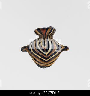 Amulett in Form eines Herzens, aus schwarzem und braunem Glas, verziert mit parallelen Linien, die auf der Vorder- und Rückseite einen Zickzack bilden, in Beige und weiß. Das Amulett ist vertikal durchbohrt, das Loch ist oben mit einem beigefarbenen Ring eingerahmt, und an den Seiten befinden sich Restgriffe. 1550–1295 v. Chr.; Neues Königreich, 18. Dynastie, Ägypten. Amulett; Höhe 1,6 cm, Breite 1,7 cm., Ein kleines Objekt mit einer runden, bauchigen Form und einem schmalen Hals, das konzentrische, wellige Bänder in schwarzen, braunen, braunen, weißen und rötlichen Tönen aufweist; die Oberfläche erscheint glatt und leicht glänzend, wobei die Bänder einen bilden Stockfoto