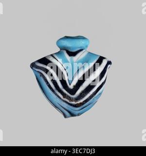 Amulett in Form eines Herzens aus blauem Glas, verziert mit parallelen Linien, die vorne und hinten einen Zickzack bilden, in Marineblau und weiß. Das Amulett ist senkrecht durchbohrt, das Loch oben mit einem blauen Ring umrahmt, an den Seiten verbleibende Griffe. 1550–1295 v. Chr.; Neues Königreich, 18. Dynastie, Ägypten. Amulett Heart/IB, Höhe 1,9 cm, Breite 1,9 cm., Ein kleines Objekt mit einer schildähnlichen Form, zentriert auf einem glatten hellgrauen Hintergrund, das geschwungene Bänder in hellem Türkis, tiefem Schwarz und weiß zeigt, die ein Chevron-Muster bilden, mit einem abgerundeten türkisfarbenen oberen Abschnitt; die Oberfläche zeigt Stockfoto