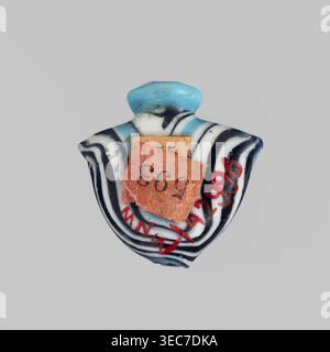 Amulett in Form eines Herzens aus blauem Glas, verziert mit parallelen Linien, die vorne und hinten einen Zickzack bilden, in Marineblau und weiß. Das Amulett ist vertikal durchbohrt, das Loch ist oben mit einem blauen Ring eingerahmt, und an den Seiten befinden sich Restgriffe. 1550–1295 v. Chr.; Neues Königreich, 18. Dynastie, Ägypten. Amulett Heart IB, Höhe 1,9 cm, Breite 1,9 cm., Ein kleines dekoratives Objekt mit einer abgerundeten Spitze in Hellblau sitzt auf einem glatten grauen Hintergrund; der Körper des Objekts zeigt abwechselnd gekrümmte Bänder in Schwarz und weiß, die einen zentralen Bereich mit Blau- und Weißtönen umgeben, und Stockfoto