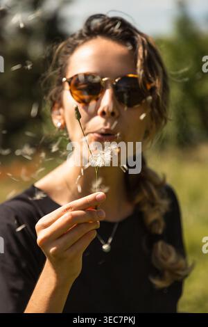 Eine junge Frau steht auf einem grünen Feld mit Sonnenbrille. Sie hält einen Löwenzahn und bläst sanft und schickt Samen in die Luft. Die Sonne scheint hell Stockfoto