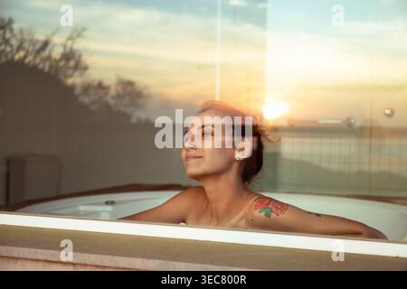 Eine junge Frau sitzt in einem Schaumbad und blickt aus dem Fenster. Sie beobachtet den Sonnenuntergang, während der Himmel seine Farbe ändert. Das Abendlicht erzeugt eine warme Atmosphäre Stockfoto