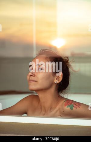Eine junge Frau sitzt in einem Schaumbad und blickt auf den Sonnenuntergang. Die Szene zeigt ihren entspannten Ausdruck, während die Sonne untergeht und Licht durch den Wind wirft Stockfoto