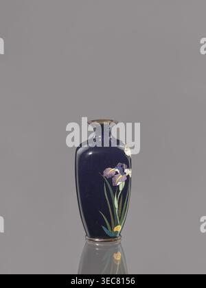 Vase, die einer ovoiden Cloisonné-Emailvase mit kurzem, dünnem, ausgestelltem Hals zugeschrieben wird; dunkelblauer Hintergrund mit Iris. Meiji-Periode, ca. 1900, Japan. Gefäß; Höhe 9,1 cm, Durchmesser 4,3 cm., Eine dunkelblaue Keramikvase mit schmalem Hals und ausgestelltem Rand sitzt mittig auf einem glatten grauen Hintergrund, auf einer Seite mit einem bemalten Blumenmotiv verziert, das lila und weiße iriähnliche Blüten, grüne lange Blätter und eine kleine gelbe Blume in der Nähe der Basis zeigt; die Vase hat ein glänzendes Finish, das Highlights und eine subtile Reflexion des Blumenmotivs auf der Oberfläche darunter. Stockfoto