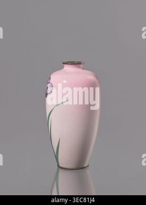 Vase, Cloisonné-Emaille, verziert mit Blumen und Iris; Taishō-Zeit, ca. 1912–1926; Nagoya, Japan; Gefäß, Höhe 15 cm, Durchmesser 7,9 cm., Eine glatte, glänzende Vase mit abgerundetem Körper und engem Hals sitzt auf einem glatten Hintergrund, die Oberfläche der Vase schattiert von blassrosa zu einem etwas tieferen Rosa nach oben. Ein partielles violettes Blütenblatt ist an der oberen Seite der Vase sichtbar, und einige lange, dünne grüne Blätter erstrecken sich entlang des Körpers nach oben. Der Rand und die Basis der Vase haben ein dünnes Metallband in einer messingähnlichen Farbe, und die Vase reflektiert die Oberfläche darunter. Die Stockfoto