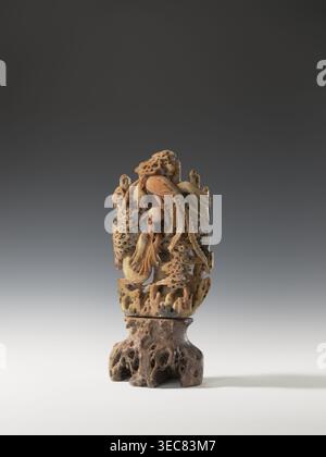 Phönix mit einem Drachen und einer Perle auf einer Basis; unbekannter Schöpfer; Chinesisch; Qing-Dynastie 1644–1911; 18. Jahrhundert; Skulptur/Figur; Material: Perlen dekorativ, Drachen mit Perle, phönix, Wolken, Felsen; Höhe 36,0 cm, Breite 17,4 cm., das Bild zeigt eine aufwendig geschnitzte vertikale Skulptur in warmen Beige- und Brauntönen, die auf einer dunkleren braunen, porösen Basis steht; der geschnitzte Abschnitt zeigt fließende organische Formen, darunter gekrümmte, federartige Elemente und abgerundete, mehrschichtige Formen mit vielen kleinen Perforationen und strukturierten Oberflächen, die ein Gefühl von Tiefe und ineinander greifenden Teilen schaffen, und die Stockfoto