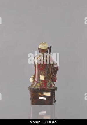 Chinesischer taoistischer unsterblicher Jap. sennin, japanische Kunst, Ikonographie der Unsterblichen, Keramikfigur eines männlichen Unsterblichen, mehrfarbige Glasur: Weiß, rot, braun, gelb, 19. Jahrhundert, Japan, möglicherweise Seto, Höhe 21,6 cm, das Bild zeigt die Rückansicht einer kleinen Figur, die auf einem rechteckigen Sockel sitzt, vor einem hellgrauen Hintergrund. Die Figur hat einen abgerundeten Kopf mit einer blassgelblichen Oberseite und dunkelbraunen Seiten und trägt ein überwiegend tiefrotes Gewand mit Flecken und Streifen aus Grün und Gelbgold. Ein Arm liegt an der Seite der Figur und hält ein blasses, kugelähnliches Objekt, der andere Stockfoto