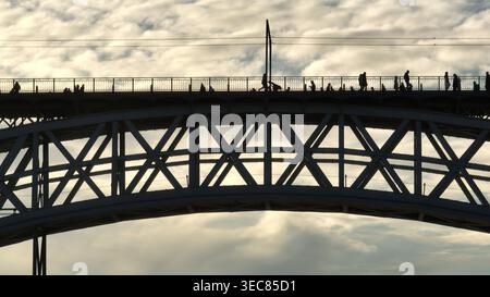 Fußgänger mit Silhouette, die bei Sonnenuntergang auf dem Oberdeck der Dom Luis I Brücke laufen, die den Fluss Douro in Porto, Portugal, überspannt und die ikonische Architektur und das geschäftige urbane Leben zeigt Stockfoto