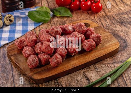 Eine Nahaufnahme roher Hackfleischbällchen in roter und rosa Farbe auf einem rustikalen Holzschneidebrett. Frisches grünes Basilikum, rote Tomaten und Wachteleier sind das Stockfoto