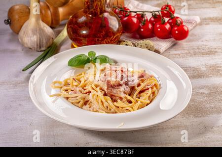 Köstliche Spaghetti Carbonara serviert auf weißem Teller, garniert mit knusprigem Speck und frischem Basilikum. Rustikaler Hintergrund mit Knoblauch, grünen Zwiebeln, Chili-Öl, Stockfoto