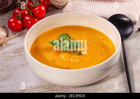 Nahaufnahme einer leuchtenden Orangencremesuppe in einer weißen Schüssel. Garniert mit frischen grünen Basilikumblättern, Kürbiskernen. Rustikaler Holzhintergrund mit rotem Chrom Stockfoto