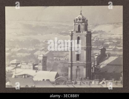 Johanniskirche; Fotografie, 1914; Thema: Panoramablick auf Vilnius einschließlich Johanniskirche; Fotograf: Jan Bułhak 1876–1950; Herkunftsort: Vilnius, Litauen; Medium: Fotografischer Druck; Bildabmessungen: 11,2 x 16,0 cm; Albumseitenabmessungen: 31,0 x 23,5 cm., Ein Graustufenfoto, das einen hohen, mehrschichtigen Turm mit bogenförmigen Öffnungen und architektonischen Details zeigt, die sich über einem Stadtbild erheben; der Turm befindet sich auf der rechten Seite des Rahmens und ist von einer Gruppe unterer Dächer und Gebäudeformen mit geneigten und Ebenen Flächen umgeben. Der Vordergrund enthält einen Cluster Stockfoto
