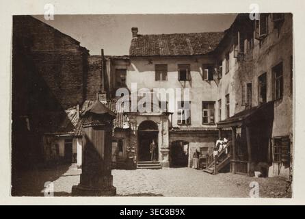 Vilnius – Ghetto. Hof im jüdischen Viertel. Fotograf: Jan Bułhak 1876–1950. 1914. Auf hellfarbenen Karton geklebt. Betrifft: Höfe, Mietshäuser in Vilnius, Litauen. Fotografischer Druck: 11,5 x 16,7 cm; Karton: 31,9 x 24,1 cm., das Bild zeigt einen alten Kopfsteinpflasterhof, der von mehrstöckigen Gebäuden mit Ziegeldächern und vielen Fenstern umgeben ist, die in Sepia-Tönen in Braun, Beige und dunkler Bräune gehalten sind; im Vordergrund steht eine verzierte Holzsäule oder Brunnenstruktur mit einem kleinen Spitzdach, die einen Schatten wirft, während ein Tor mit einem gewölbten Giebel und Stufen Stockfoto