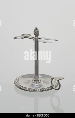 Scherenförmiger Kerzenhalter stoczek, Jan Radecki fl. 1815–1837, Goldschmied. Kerzenhalter für eine dünne Kerze, die um den Stiel geschleift war; gestützt auf eine plattenförmige Basis, die mit einem Halbzylinder umrandet wurde und mit einem Ringgriff mit Daumenauflage versehen war. Der Stiel wurde als Hülse geformt, um die die dünne Kerze Serpentine verdrehte; die Hülse wurde mit einem Kiefernzapfen und einem Schraubstock in Form einer flachen Schere mit Spiralgriffen gekrönt, um die Kerze auf die gewünschte Länge zu verlängern. 1815–1837, Poznań, Polen. Funktion: Beleuchtung. Höhe 14,5 cm; Breite 14 cm; Tiefe 11 cm; Ein Metallstativ mit rundem Fuß Stockfoto