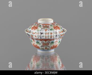 Schüssel mit Deckel; Chinesisch, Qing-Dynastie (Stil von Qianlong, 1736–1795); Porzellan mit polychromen Emails; dekoriert mit Fledermäusen, Langlebigkeit Shou, Lotusranken, Lotusblüten, und Pfirsiche; Gefäßhöhe 9 cm, Durchmesser 10,7 cm; datiert 1736–1795. Eine Porzellanschale mit Deckel, abgerundetem Körper und ausgestelltem Rand sitzt mittig auf einem grauen Hintergrund, deren glänzend weiße Oberfläche mit einem floralen und blättrigen Design in rot-orangen, grünen, blauen und gelben Akzenten verziert ist, mit wiederholten Blumenmotiven und Blättern, die Deckel und Körper bedecken; die Schale wirft eine sanfte Reflexion unten auf der reflektierenden Oberfläche; Stockfoto