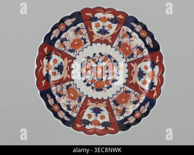 Teller, Japanisch, Meiji (1868–1912), Imari, Blumenmotive einschließlich Lotusblumen, Pfirsiche (ikonographisch) und Chrysanthemen, 19./20. Jahrhundert, Japan; Gefäß, Durchmesser 31,2 cm., Eine runde dekorative Platte mit einem gewellten Rand, von oben auf einem grauen Hintergrund dargestellt, mit einem symmetrischen Blumenmuster in Rot, Blau, weiß und Orange; ein zentrales kreisförmiges Medaillon aus Blumenmotiven wird von einem strukturierten weißen Ring umgeben, dann abwechselnd Platten mit roten gewellten Abschnitten und blau umrandeten Blumenabschnitten, die alle mit stilisierten Blumen und Blättern in Rot, Orange und Blau gefüllt sind Stockfoto