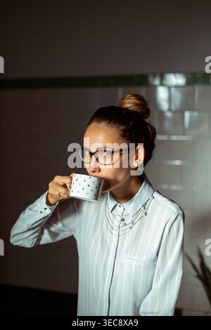 Eine junge Frau mit einem Brötchen trinkt aus einer kleinen Tasse in einem dunklen Zimmer. Sie steht in der Nähe eines Fensters, das weiches Licht hereinlässt. Die Einstellung verfügt über ein einfaches und q Stockfoto