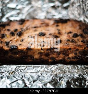 Nahaufnahme eines in Scheiben geschnittenen hausgemachten Brotpuddings, gefüllt mit getrockneten Früchten, die auf zerknitterter Folie ruhen, zeigt die reiche goldene Kruste und die dichte, feuchte Textur o Stockfoto