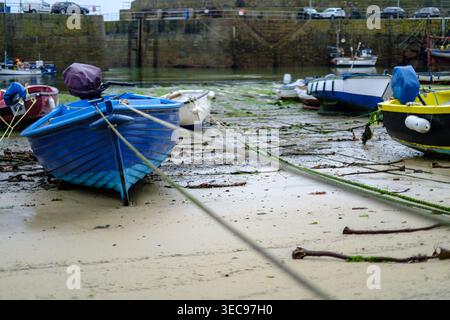 Farbenfrohe Fischerboote liegen bei Ebbe auf dem sandigen Hafenboden in Mousehole, Cornwall, England, mit Seilen, die sich in Richtung des Steins erstrecken Stockfoto
