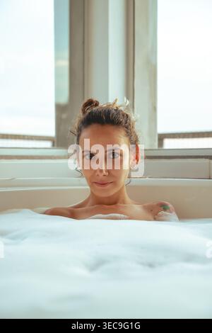 Eine junge Frau mit gebundenen Haaren entspannt sich in einem Schaumbad. Sie sitzt in einer weißen Wanne, umgeben von Blasen. Licht kommt durch das Fenster, während sie ihn genießt Stockfoto