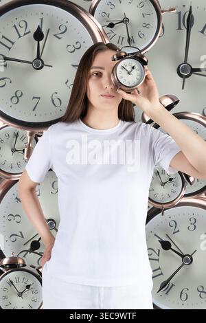 Frau mit Wecker, die in die Kamera schaut, umgeben von vielen Uhrengesichtern Stockfoto