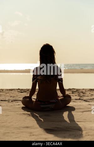 Eine junge Frau sitzt in Lotusposition am Strand und trägt einen Bikini. Sie meditiert, während die Sonne über dem Meer untergeht. Der Himmel ist hell orange und das W Stockfoto
