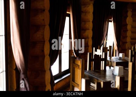Blick auf ein gemütliches, leeres rustikales Restaurant mit Holzwänden, dunklen Vorhängen, Holztischen und Stühlen. Helle Fenster beleuchten den Raum, h Stockfoto
