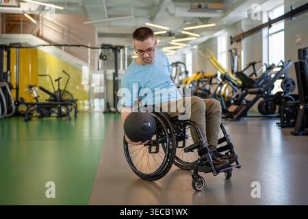 Mann-Athlet im Rollstuhl mit Medizinball zum Aufbauen der Armstärke bei der posttraumatischen Sitzung im Fitnessstudio. Stockfoto