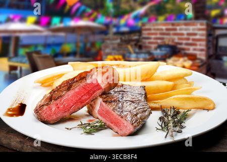 Gegrilltes Picanha-Steak, mittelselten mit dicken Pommes frites auf einem weißen Teller serviert, mit einem verschwommenen traditionellen brasilianischen Festa Junina Backgrou Stockfoto