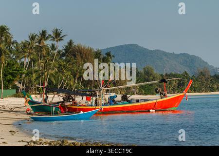 Farbenfrohe traditionelle thailändische Longtail-Boote liegen in der Nähe eines tropischen Sandstrandes mit Palmen und Hügeln, ruhige Küstenlandschaft in Thailand Stockfoto