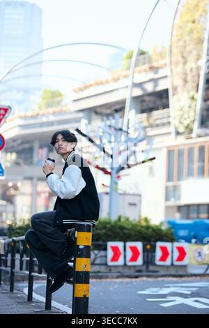 Stilvoller japanischer junger Mann, der auf einer Leitplanke sitzt und Kaffee trinkt und in der Stadt in die Kamera schaut Stockfoto