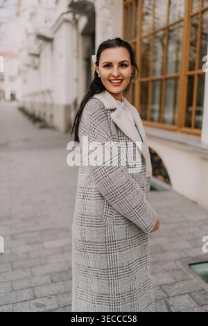Frau Mantel urbane lächelnde junge Frau in einem grauen karierten Mantel spaziert an einem sonnigen Tag auf einem Bürgersteig der Stadt Stockfoto