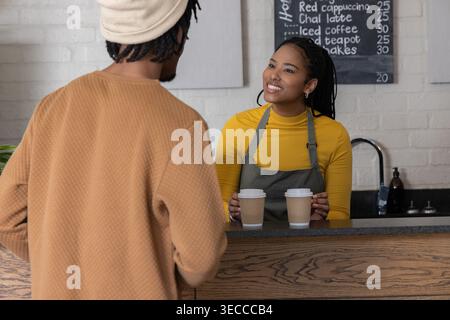 Afroamerikanischer Barista mit Schürze überreicht männlichen Gästen am Café-Theke zwei To-go-Kaffeetassen Stockfoto