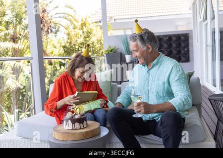 Seniorenpaar mit Partyhüten, Goldgeschenk auf der Terrasse öffnen und Wein um 50 Kuchen halten. Partner, Geburtstag, Outdoor, Balkon, Geschenk, entpacken, Stockfoto